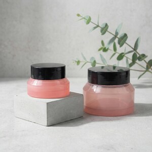Pode incluir: Dois frascos transparentes com tampas pretas, cheios de uma substância rosa, são exibidos. Um frasco está sobre um pequeno bloco retangular cinza. Folhas verdes estão no fundo. Os frascos são provavelmente para cosméticos ou produtos para a pele.