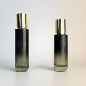 May include: Two cylindrical glass bottles with a gradient black to clear design. Each bottle has a gold-colored cap. The bottles are standing upright against a white background. The bottles are likely for perfume or essential oils.
