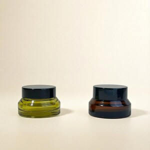 May include: Two small, round glass jars with black lids. One jar is olive green, the other is amber brown. The jars are on a light beige surface. The jars are likely for storing cosmetics or other small items.