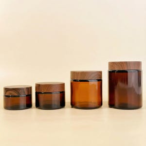 May include: Four amber glass jars with wooden lids of different sizes are arranged in a row. The jars have a cylindrical shape and are made of a translucent brown glass. The wooden lids provide a natural contrast to the glass.