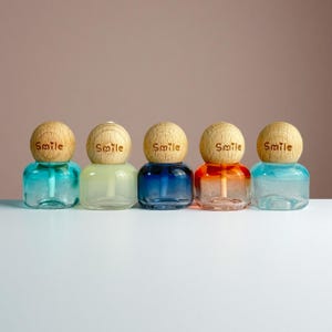 Könnte beinhalten: Fünf kleine, farbige Glasflaschen mit kugelförmigen Holzverschlüssen. Jede Flasche enthält eine Flüssigkeit in einer anderen Farbe: Türkis, Hellgrün, Dunkelblau, Orange und Hellblau. Das Wort "Smile" ist auf jeden Holzverschluss geätzt.