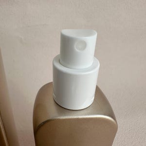 May include: Close-up of a gold-colored bottle with a white spray nozzle. The nozzle has a rounded top with a small opening. The bottle has a smooth, curved shape.