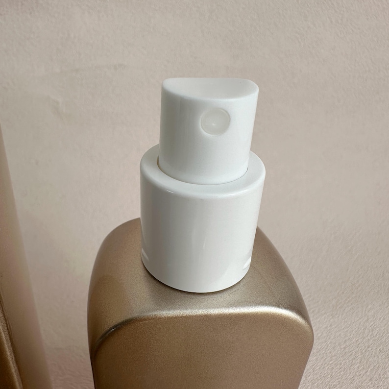 May include: Close-up of a gold-colored bottle with a white spray nozzle. The nozzle has a rounded top with a small opening. The bottle has a smooth, curved shape.