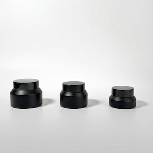 Könnte beinhalten: Drei schwarze Kosmetikgläser mit passenden schwarzen Deckeln sind in einer Reihe angeordnet. Die Gläser sind zylindrisch mit einem etwas breiteren Boden und einer glatten, matten Oberfläche. Die Deckel sind flach und kreisförmig.