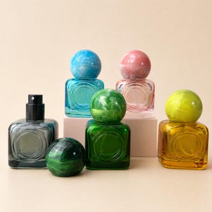 Op de afbeelding: Een collectie van vijf vierkante glazen parfumflesjes met bolvormige doppen. De flesjes zijn in de kleuren blauw, roze, groen, geel en donkergrijs. Elk flesje heeft een cirkelvormige inkeping aan de voorzijde en een bijpassende gemarmerde bolvormige deksel.