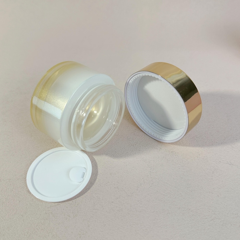 May include: An open, clear glass jar with a gold-colored rim and a white interior. The jar is partially frosted with a gold metallic accent. The lid is off, revealing the inside of the jar. A small white lid is next to the jar.