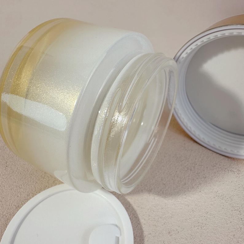 May include: A clear glass jar with a white lid and a gold-colored rim. The jar is open, revealing a white substance inside. The lid is off to the side. The jar is tilted, and the contents are partially visible.