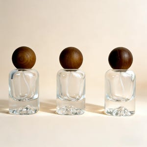 May include: Three clear glass perfume bottles with wooden spherical caps. The bottles have a rectangular shape with a rounded base. The wooden caps are a dark brown color. The background is a solid, light beige color.