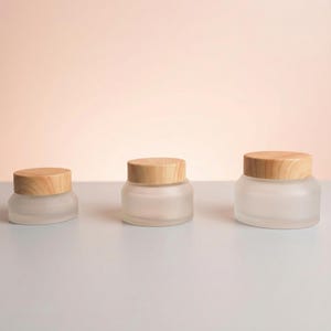 Pode incluir: Três frascos de vidro fosco com tampas de madeira de tamanhos variados. Os frascos estão dispostos em uma fileira, mostrando um design minimalista. As tampas são de madeira clara, contrastando com o vidro fosco. O fundo é de cor neutra.