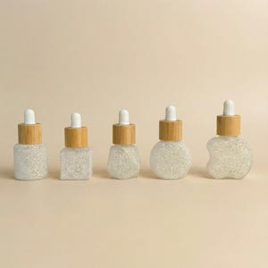 May include: Five clear glass dropper bottles with white and silver glitter inside. Each bottle has a different shape and a bamboo-colored cap with a white dropper. The bottles are arranged on a light beige background.