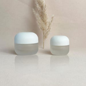 May include: Two small, frosted glass jars with rounded white lids. The jars are displayed on a light-colored surface, with a dried plant stem in the background. The jars are likely for cosmetics or skincare products.
