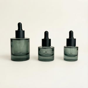 May include: Three glass dropper bottles in varying sizes. The bottles are a smoky gray color with black droppers. The bottles are cylindrical with a rounded base. The background is a solid, neutral color.