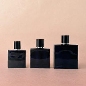 Op de afbeelding: Drie donkerblauwe rechthoekige parfumflessen met zwarte doppen en zilveren kragen worden gepresenteerd tegen een lichtroze achtergrond. De flessen variëren in grootte, waardoor een visueel aantrekkelijke opstelling ontstaat.