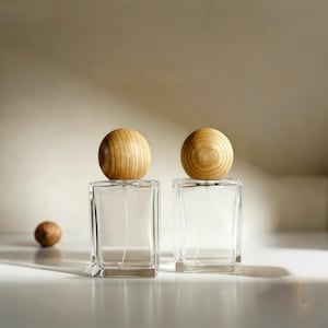 Puede incluir: Dos frascos de perfume de vidrio transparente con tapones esféricos de madera. Los frascos rectangulares se asientan sobre una superficie blanca. Una sola esfera de madera está a la izquierda de los frascos. El fondo es de color neutro.