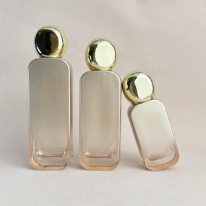 May include: Three rectangular perfume bottles with rounded edges and gold-colored caps. The bottles have a gradient color scheme, transitioning from a light beige to a darker beige. One bottle is tilted to the side.