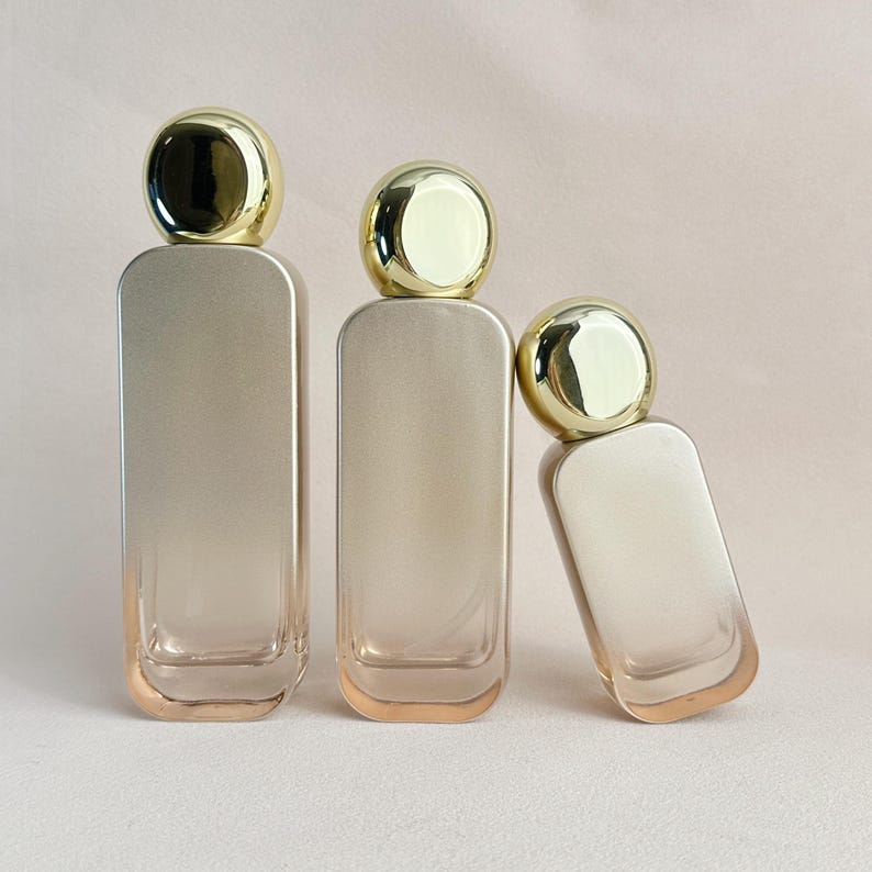 May include: Three rectangular perfume bottles with rounded edges and gold-colored caps. The bottles have a gradient color scheme, transitioning from a light beige to a darker beige. One bottle is tilted to the side.