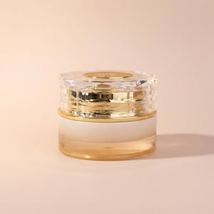 Puede incluir: Un tarro cosmético transparente y dorado con tapa facetada. El tarro tiene una banda dorada en el medio y una parte superior dorada. El producto es probablemente una crema o loción, y la estética general es lujosa y elegante.