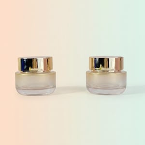 May include: Two empty cosmetic jars with frosted glass bases and reflective gold-toned screw-on lids. The jars feature a cylindrical shape with a smooth, rounded bottom edge, showcasing a clear glass construction that transitions to a frosted finish.