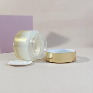 May include: A clear glass jar with a gold-colored lid and a small white lid. The jar has a frosted appearance with a gold gradient on the side. The lid is off to the side.