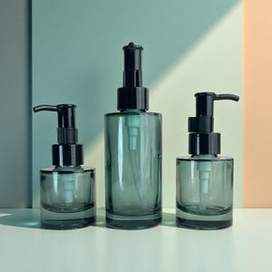 May include: Three glass soap or lotion dispensers in varying sizes. The bottles are a muted teal color with black pump tops. The dispensers are arranged on a white surface against a two-tone teal and peach background.