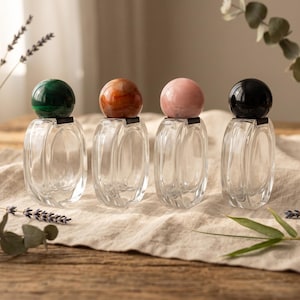 Op de afbeelding: Vier doorzichtige glazen parfumflesjes met ovale lichamen en bolvormige doppen in groen, amber, lichtroze en zwart. Elk flesje heeft een glad, transparant ontwerp met een opvallende gekleurde bolvormige dop, gerangschikt in een rij.