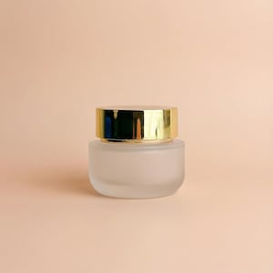 Op de afbeelding: Een klein, matglazen potje met een goudkleurige deksel. Het potje heeft een afgeronde vorm en staat op een lichtperzikkleurige achtergrond. De deksel reflecteert licht, waardoor het glanzend lijkt.