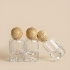 May include: Three clear glass spray bottles with wooden spherical caps. The bottles have a rounded base and a spray nozzle. One bottle is tilted to the side, while the others stand upright against a neutral background.