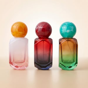 May include: Three perfume bottles with spherical caps in orange, red, and teal. The bottles have a geometric design and a gradient color scheme, transitioning from light to dark. The bottles are displayed on a neutral background.