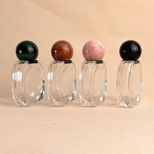 May include: Four clear glass perfume bottles with decorative, spherical stoppers in green, brown, pink, and black. The bottles have an oval shape and sit on a neutral background.