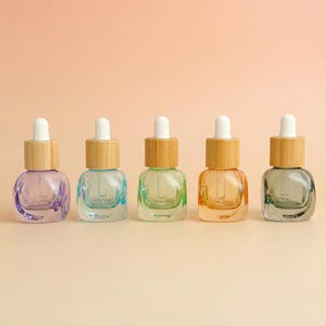 Könnte beinhalten: Fünf Glas-Tropfflaschen mit Holzdeckeln und weißen Tropfern, jede mit einer andersfarbigen Flüssigkeit gefüllt: lila, blau, grün, orange und grau. Die Flaschen stehen auf einem pfirsichfarbenen Hintergrund.