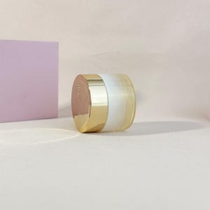 May include: A cosmetic jar with a gold-colored lid and base, and a white and yellow-tinted glass body. The jar is cylindrical and sits on a light-colored surface. A light purple square is in the background.