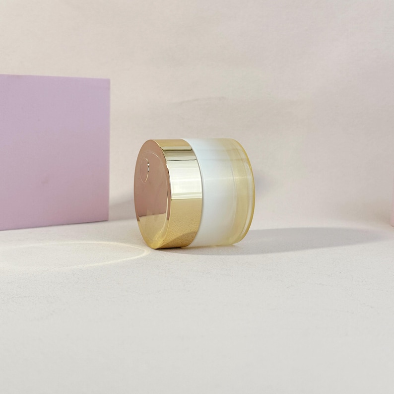 May include: A cosmetic jar with a gold-colored lid and base, and a white and yellow-tinted glass body. The jar is cylindrical and sits on a light-colored surface. A light purple square is in the background.