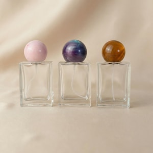 May include: Three clear glass perfume bottles with decorative spherical stoppers. The stoppers are pink, purple and blue, and brown. The bottles are rectangular with a spray nozzle inside. The background is a solid beige color.