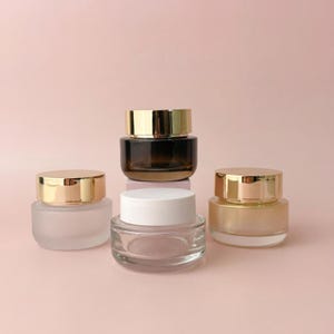 Pode incluir: Quatro pequenos frascos de vidro com tampas douradas. Um frasco é fosco, um é transparente, um é âmbar e um é de cor âmbar claro. Os frascos estão dispostos sobre um fundo rosa.