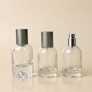 May include: Three clear glass spray bottles with cylindrical bodies and thick bases. Two bottles feature matte grey caps, while the third bottle on the right has a silver spray nozzle mechanism exposed without a cap. Each bottle includes a clear dip tube.