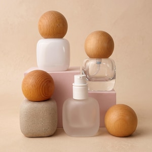 May include: A collection of cosmetic containers featuring square-shaped bottles and jars with rounded wooden sphere caps. The set includes a white opaque bottle, a clear glass bottle with a pump, a frosted glass bottle, and a speckled beige jar.