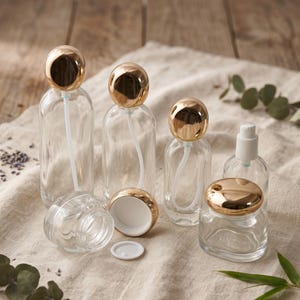 Könnte beinhalten: Eine Sammlung von klaren Glasbehältern, bestehend aus drei hohen Flaschen mit goldenen kugelförmigen Verschlüssen und weißen Pumpenröhrchen, einem kleinen Tiegel mit goldenem Deckel sowie einem kleinen Tiegel mit goldenem Verschluss und weißer Dichtscheibe.