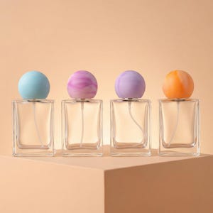Puede incluir: Cuatro frascos de perfume de vidrio transparente con tapones esféricos de colores. Los tapones son en tonos azul, rosa, morado y naranja. Los frascos son rectangulares y están sobre una superficie marrón claro.