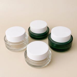 May include: Four small glass jars with white lids. Two jars are clear, and two are a dark green color. The jars are round with a low profile, and the lids are flat and circular. The jars are arranged on a light beige surface.
