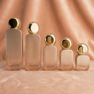 May include: Five perfume bottles of varying sizes are arranged in a row. Each bottle has a rectangular body with rounded corners and a gold-colored, spherical cap. The bottles have a soft, neutral color, and the background is a matching fabric.
