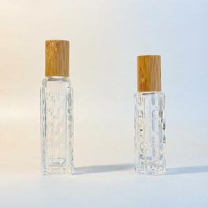 May include: Two clear glass roller bottles with wooden caps. The bottles have textured patterns on the glass. One bottle has a diamond pattern, and the other has a vertical, organic pattern. The wooden caps are light brown.
