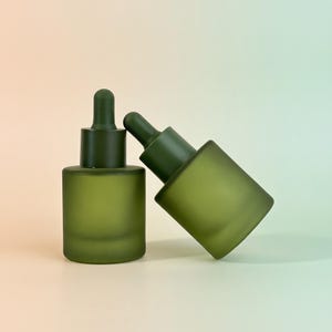 Könnte beinhalten: Zwei zylindrische Glastropfflaschen mit mattem olivgrünem Finish. Eine Flasche steht aufrecht, während die andere in einem Winkel geneigt ist und sich an die erste anlehnt. Beide verfügen über passende dunkelgrüne Tropfverschlüsse mit integriertem Ball.