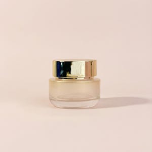 Op de afbeelding: Een kleine, heldere glazen pot met een matte afwerking, afgedekt met een glanzend goudkleurig deksel. De pot staat op een lichtroze oppervlak, wat een minimalistische esthetiek creëert. De pot is waarschijnlijk voor cosmetica of huidverzorgingsproducten.