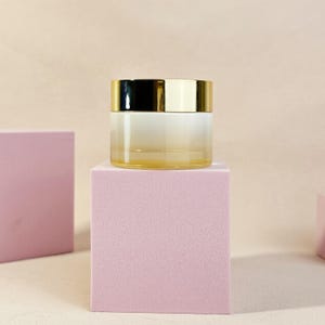 May include: A small, clear glass jar with a gold-colored lid. The jar is sitting on a pink cube. The jar has a gradient effect, transitioning from a light yellow to white. The background is a light beige color.