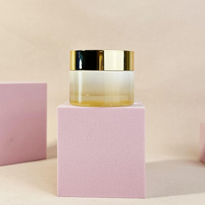 May include: A small, clear glass jar with a gold-colored lid. The jar is sitting on a pink cube. The jar has a gradient effect, transitioning from a light yellow to white. The background is a light beige color.