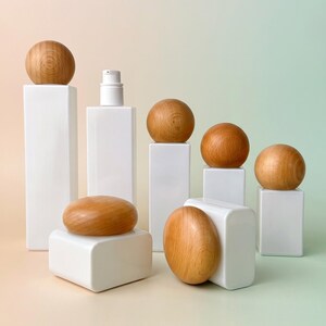 Op de afbeelding: Een collectie witte rechthoekige cosmetische verpakkingen van verschillende hoogtes, elk voorzien van een gladde, bolvormige houten dop. Eén verpakking heeft een witte pompdispenser, terwijl andere zijn afgewerkt met houten bollen.