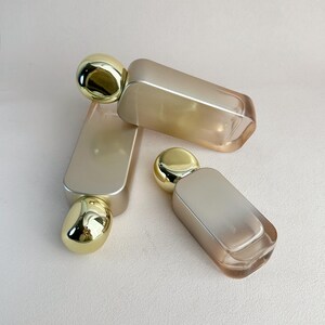 May include: Three perfume bottles with a rectangular shape and rounded gold caps. The bottles have a gradient color scheme, transitioning from a light gold to a soft, translucent hue. The bottles are arranged on a neutral background.