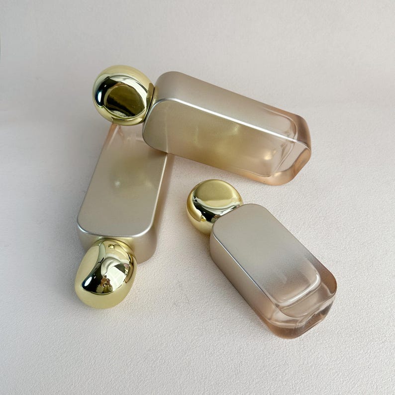 May include: Three perfume bottles with a rectangular shape and rounded gold caps. The bottles have a gradient color scheme, transitioning from a light gold to a soft, translucent hue. The bottles are arranged on a neutral background.