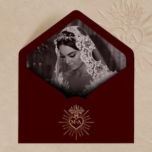 Puede incluir: Una invitación de boda burdeos con una foto en blanco y negro de una novia con velo de encaje. El sobre tiene un emblema dorado con las iniciales "M-A" y un diseño de corazón. El fondo es de color beige claro.