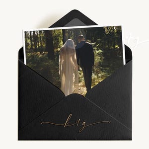 Pode incluir: Um envelope preto com uma fotografia de um casal caminhando em uma floresta. A fotografia é visível dentro do envelope. O envelope tem uma inscrição dourada das letras "k+g" na frente.